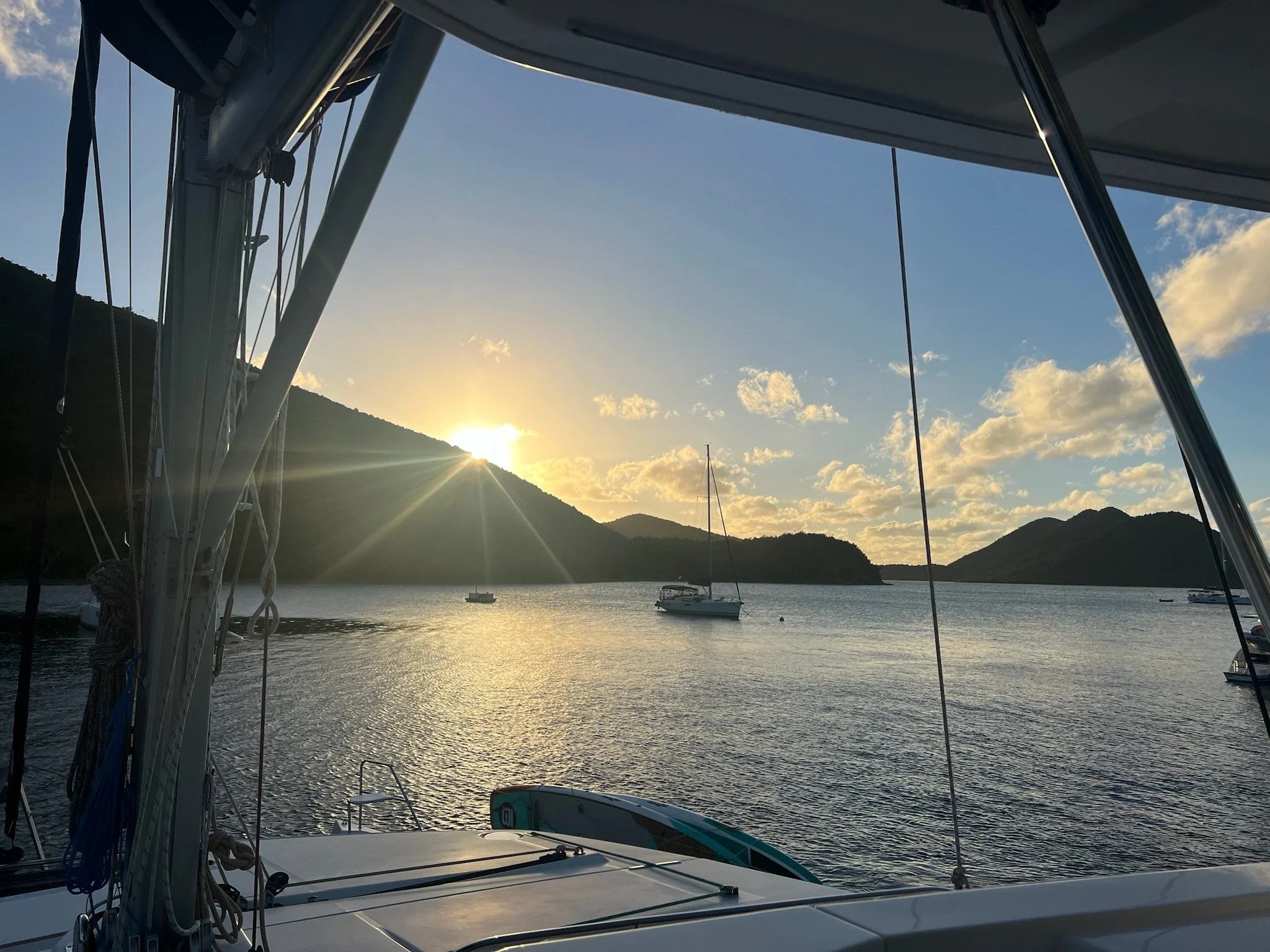 Sunset over a calm body of water viewed from a boat with visible rigging and parts of the boat in the foreground. Several sailboats are anchored in the distance near hilly shoreline.