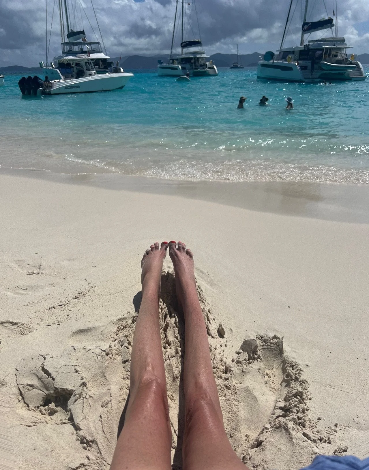 Person relaxing on a sandy beach with their legs extended, facing turquoise water with boats moored, people swimming, and distant mountains under cloudy sky.