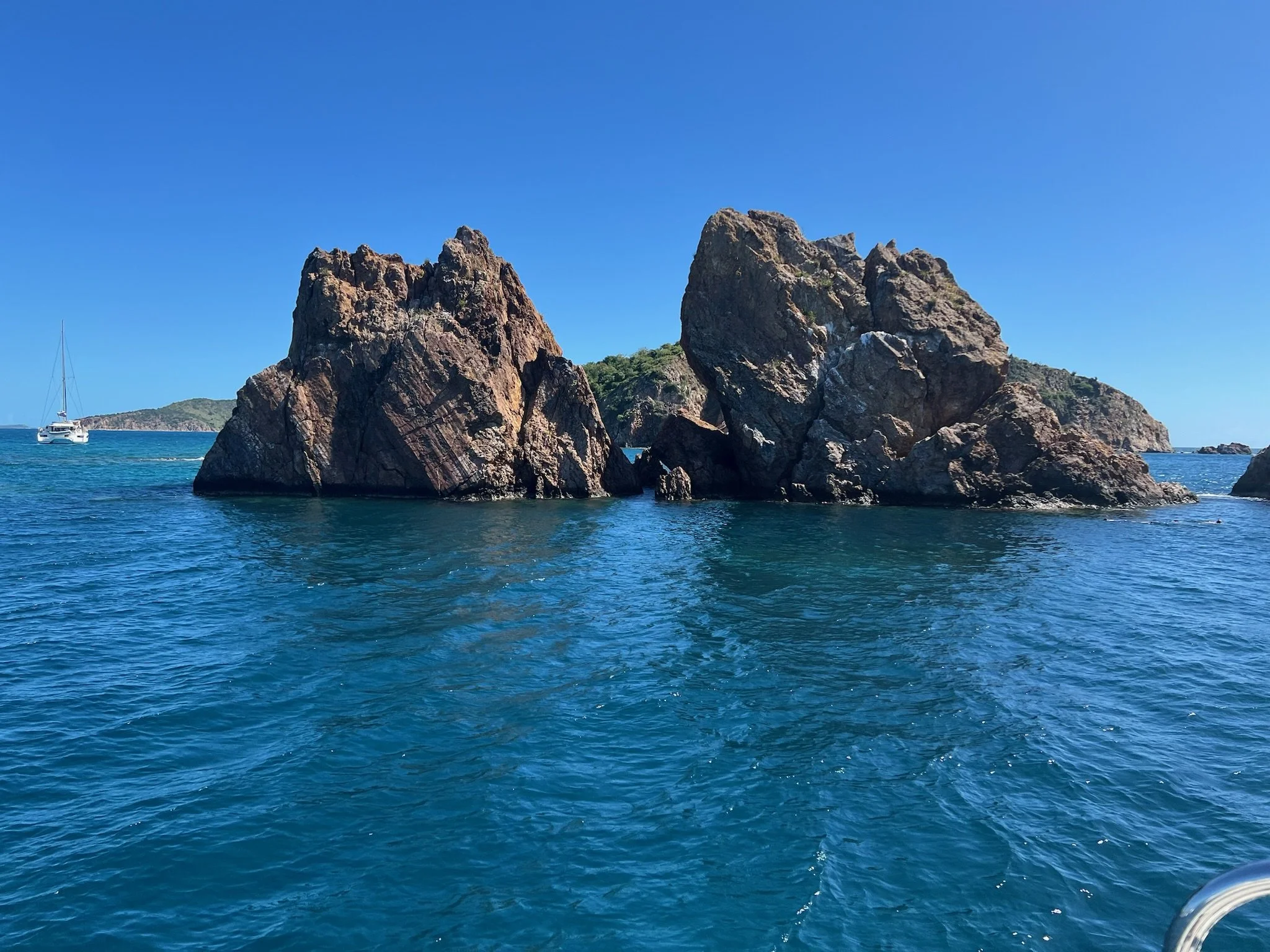 Two large rocky formations in the ocean with clear blue water, a sailboat in the distance, and a small island or landmass on the horizon under a bright blue sky.