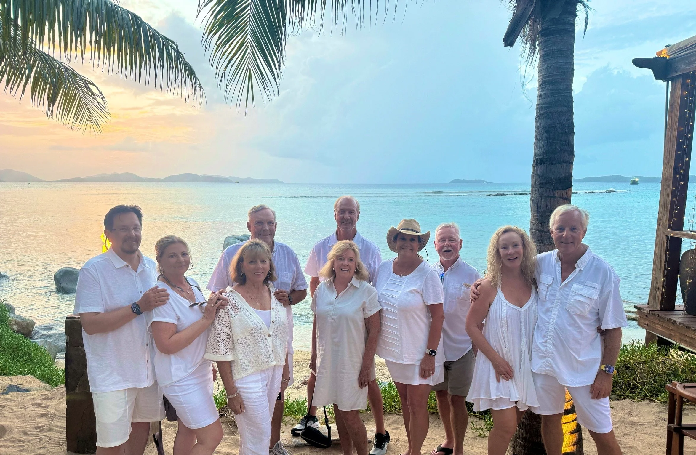 Group of twelve people in white clothing posing on a sandy beach with the ocean and islands in the background during sunset.