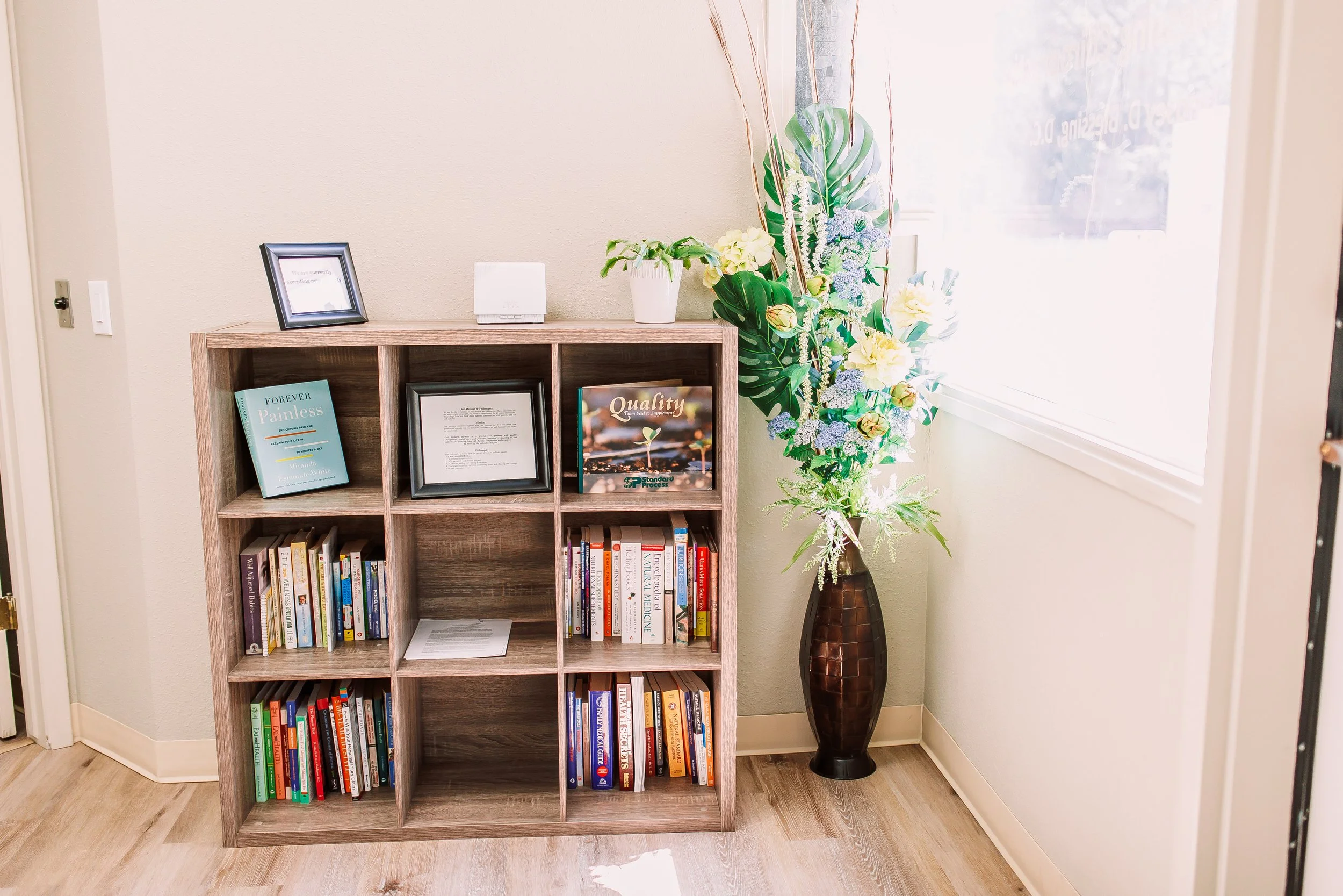 A wooden bookshelf with books and framed documents, a potted plant, and a floral arrangement in a tall vase, situated near a window in a room.