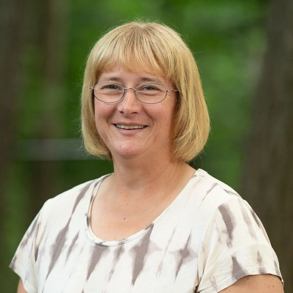 A smiling woman with blond hair, glasses, and a tie-dye shirt standing outdoors with blurred green trees in the background.
