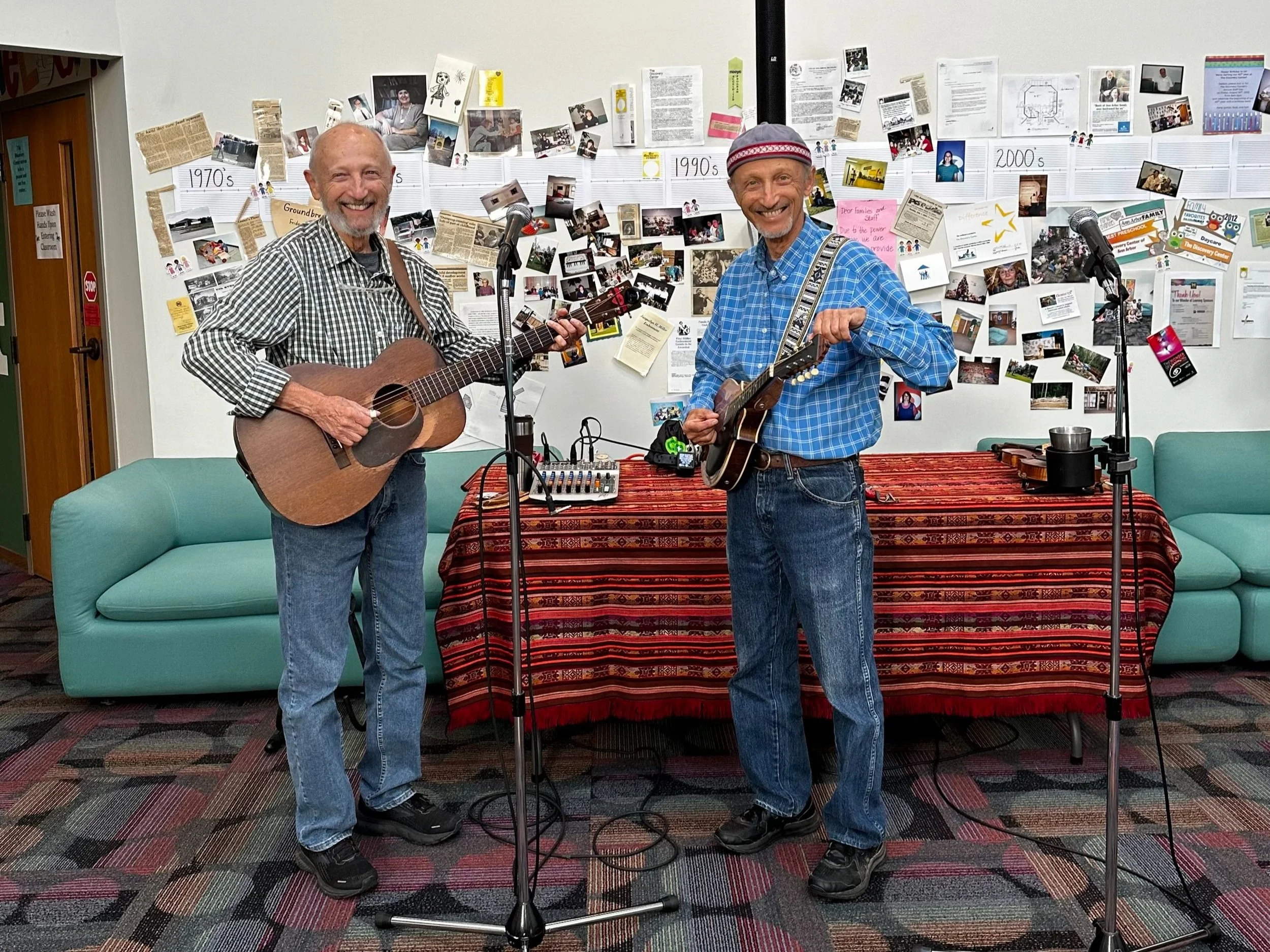 Two men playing string instruments in a room with a collage of photos and papers on the wall. One man is playing a guitar, the other a mandolin. There are microphones in front of them.
