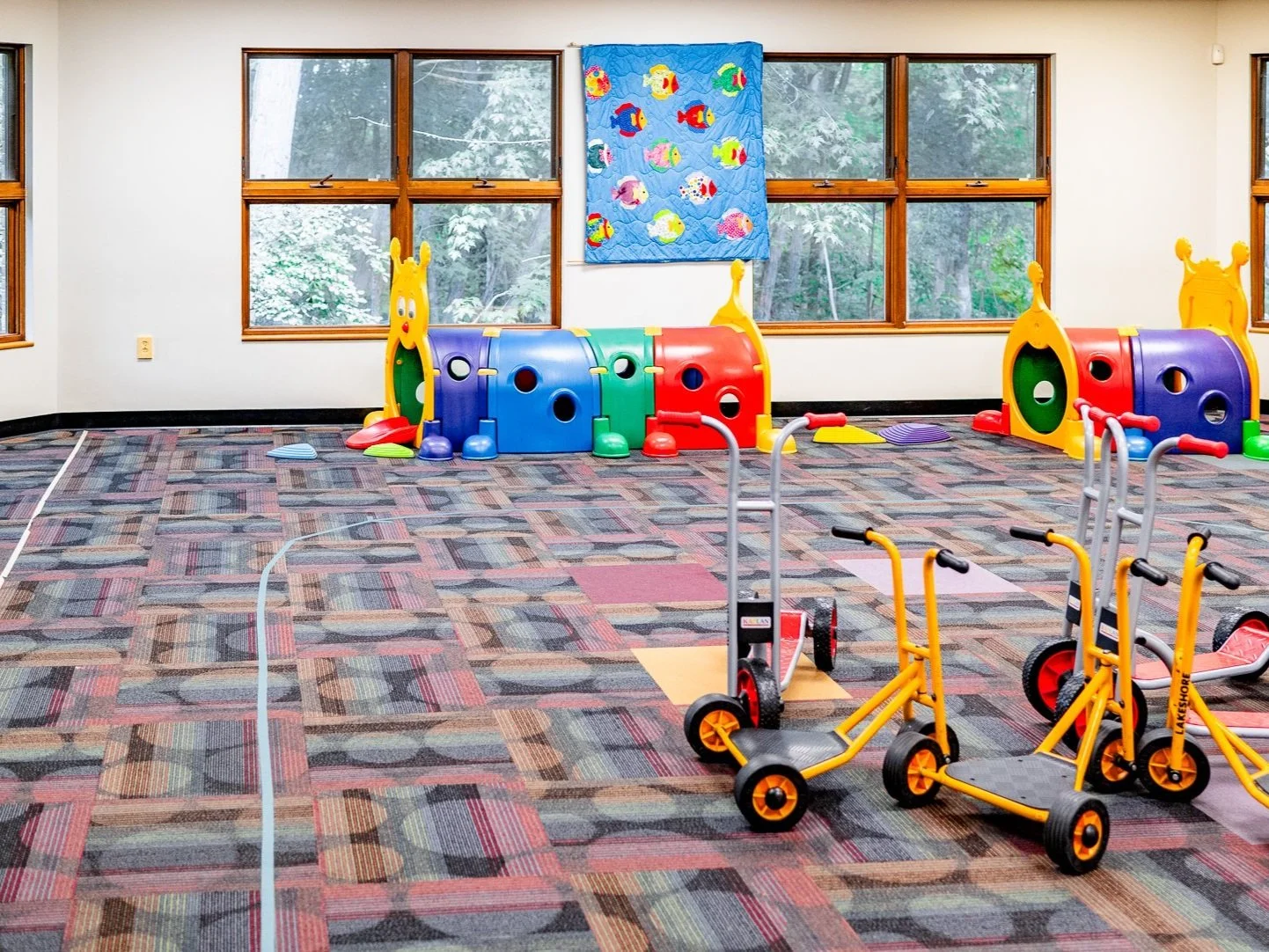 Indoor playroom with colorful plastic play tunnels, scooters, and a patterned carpet floor.