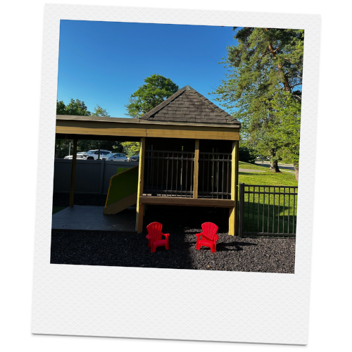 Backyard playhouse with red plastic chairs, surrounded by trees and grass on a sunny day.