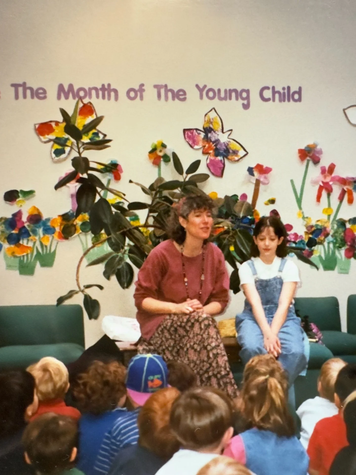 A woman and a girl sitting on a bench in front of a colorful wall with butterflies and flowers. The wall features the text 'The Month of The Young Child'. The audience, consisting of children, is seated on the floor in front of them.