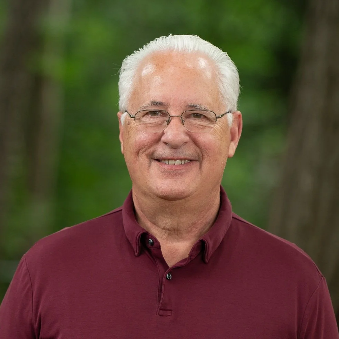 A smiling elderly man with white hair and glasses, wearing a maroon button-up shirt, standing outdoors with a blurred green background.