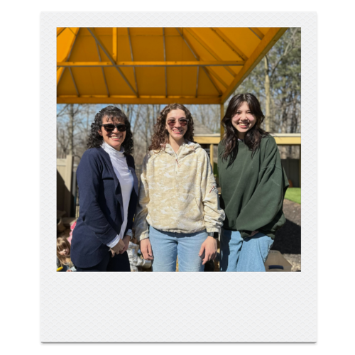 Three young women smiling outdoors under a yellow canopy.
