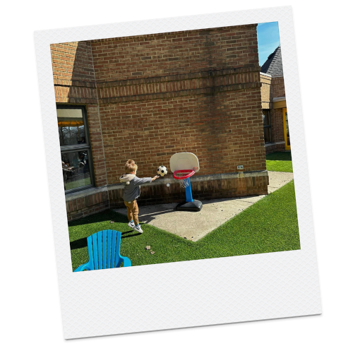 A young boy playing with a soccer ball on a small concrete patio outside a house, with a portable basketball hoop nearby.