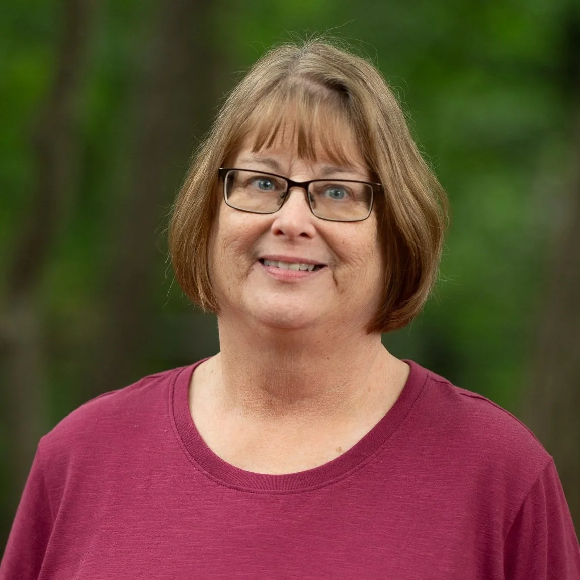 A woman with glasses and short brown hair, wearing a maroon shirt, smiling outdoors with a blurred green forest background.
