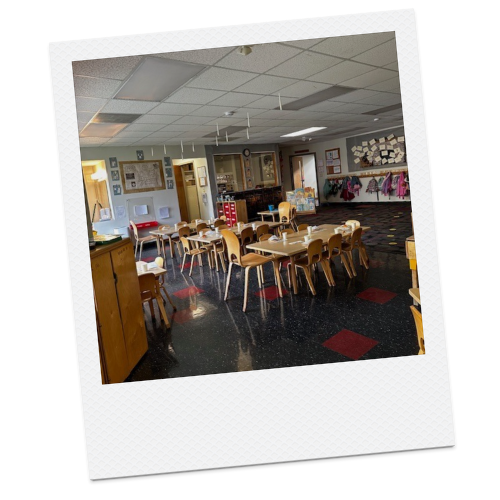 A classroom or daycare room with small tables and chairs, bulletin boards, and bright decorations.
