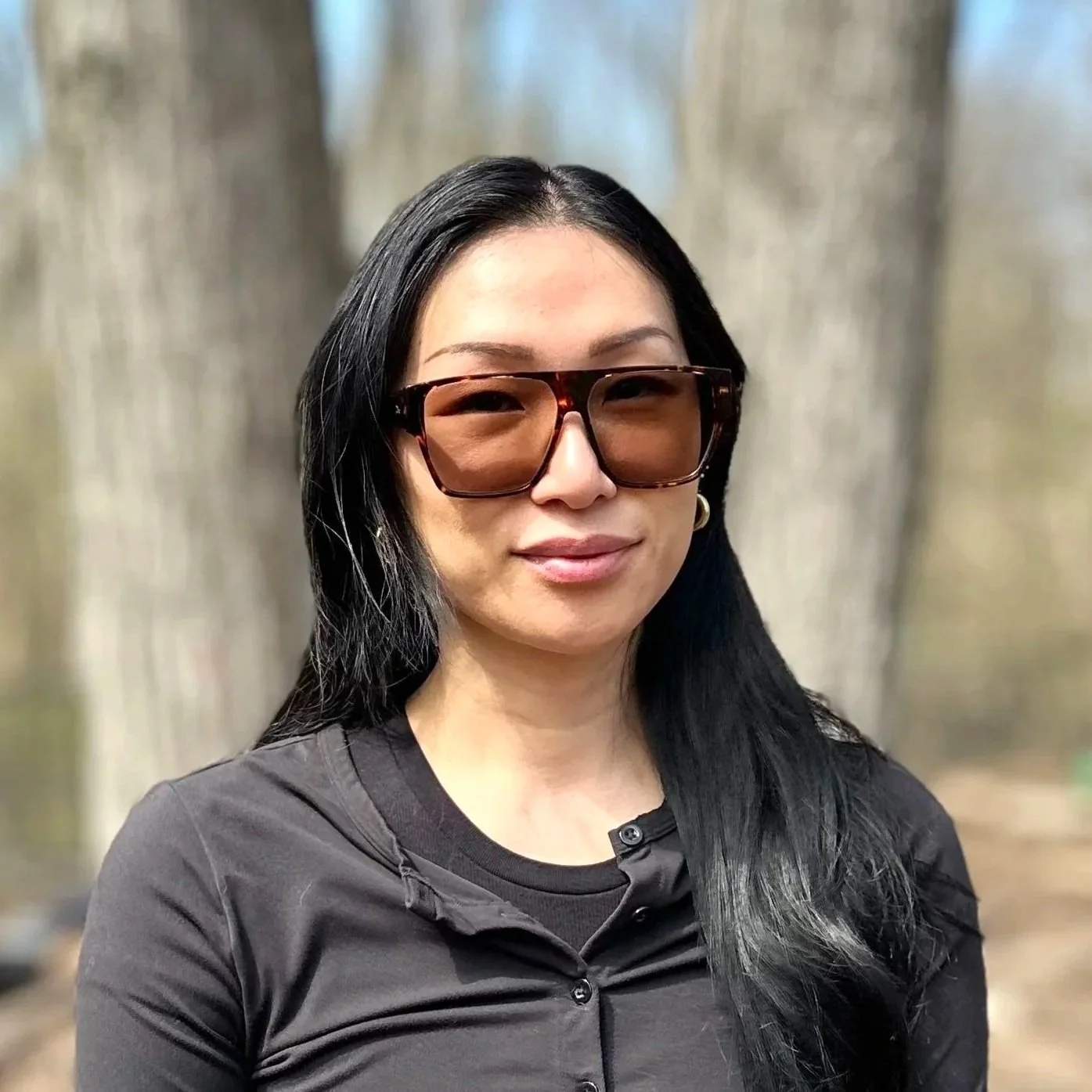 A woman with long black hair wearing sunglasses and a black shirt, standing outdoors with trees in the background.