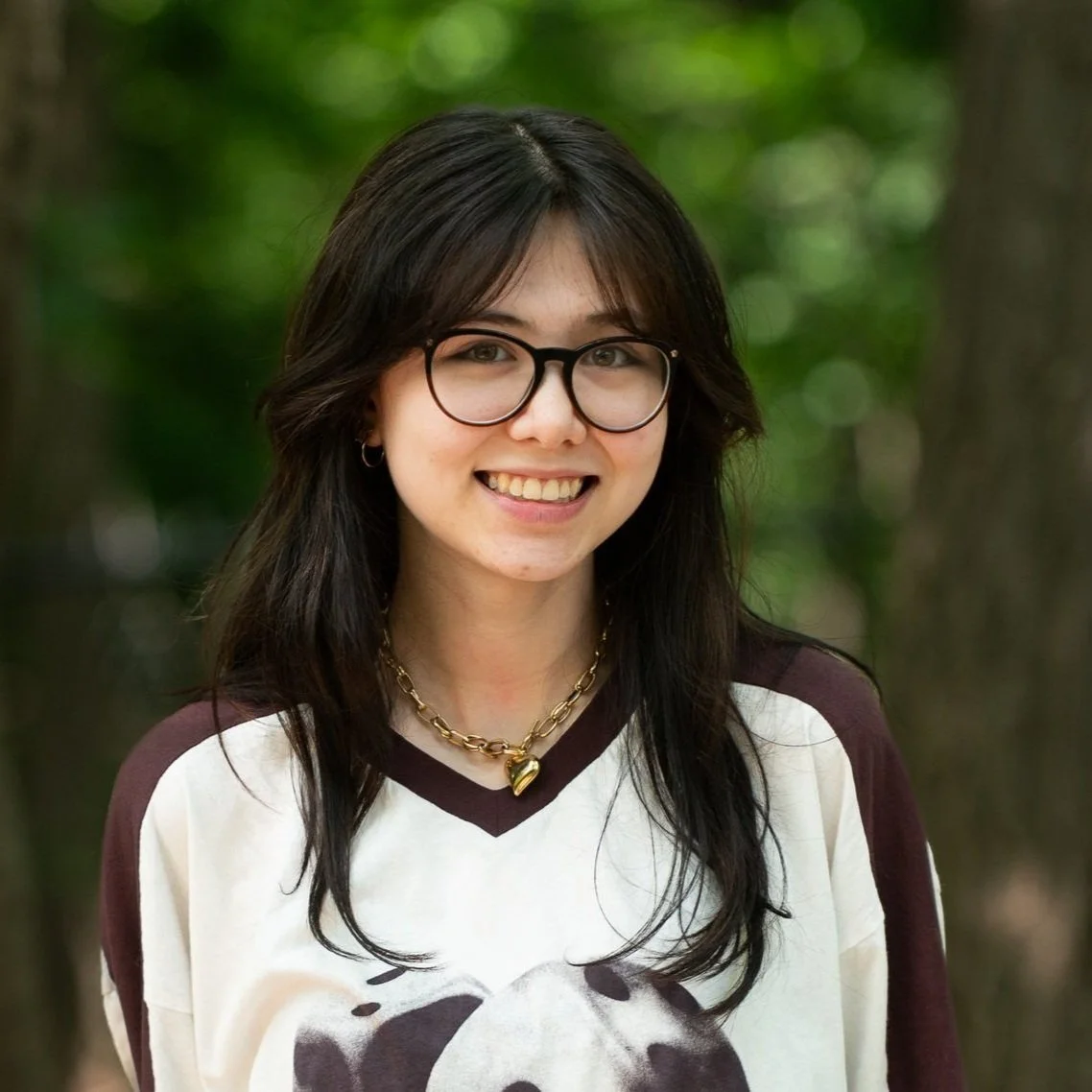 Young woman with glasses smiling outdoors, wearing a panda graphic sweatshirt and a gold chain necklace with a heart pendant, with a forest background.
