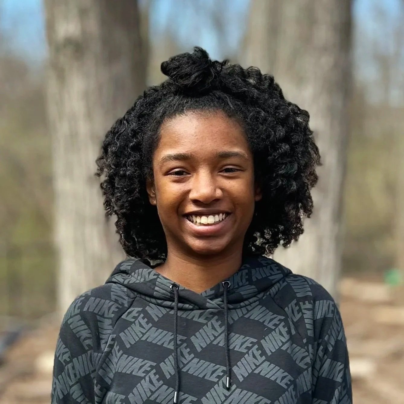 A young woman with curly black hair, smiling outdoors with blurred trees in the background, wearing a black Nike hoodie.