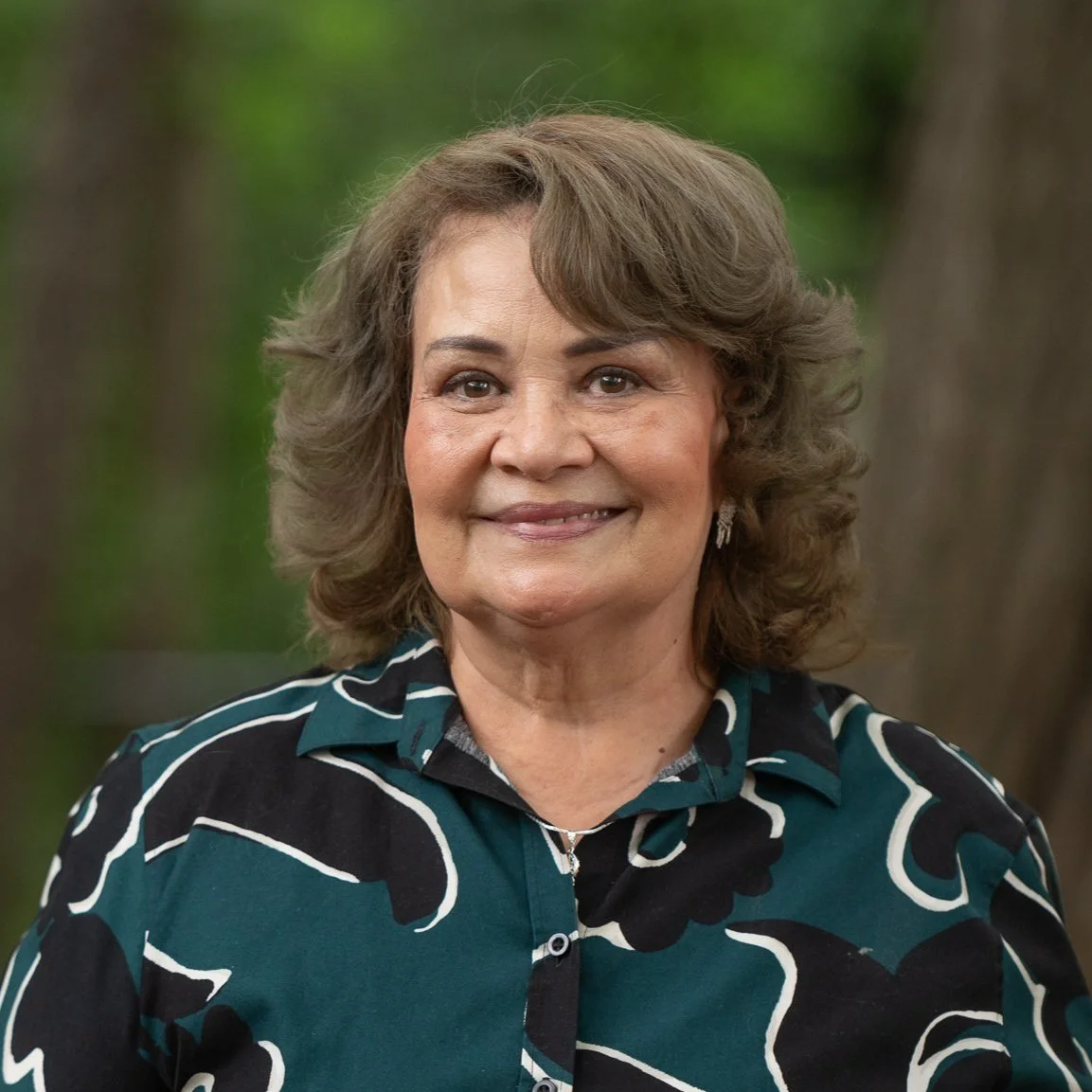A smiling middle-aged woman with curly brown hair, wearing a black shirt with white and green patterns, standing outdoors with blurred greenery in the background.