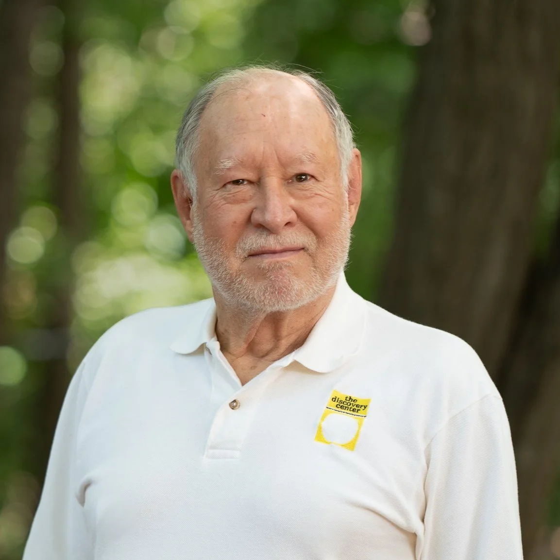 Older man with gray hair and beard wearing a white polo shirt with a yellow logo, standing outdoors in a wooded area.