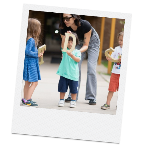 A woman and three young children outdoors, with the woman helping a girl hold a wooden horseshoe, while two other children hold plush horseshoes.