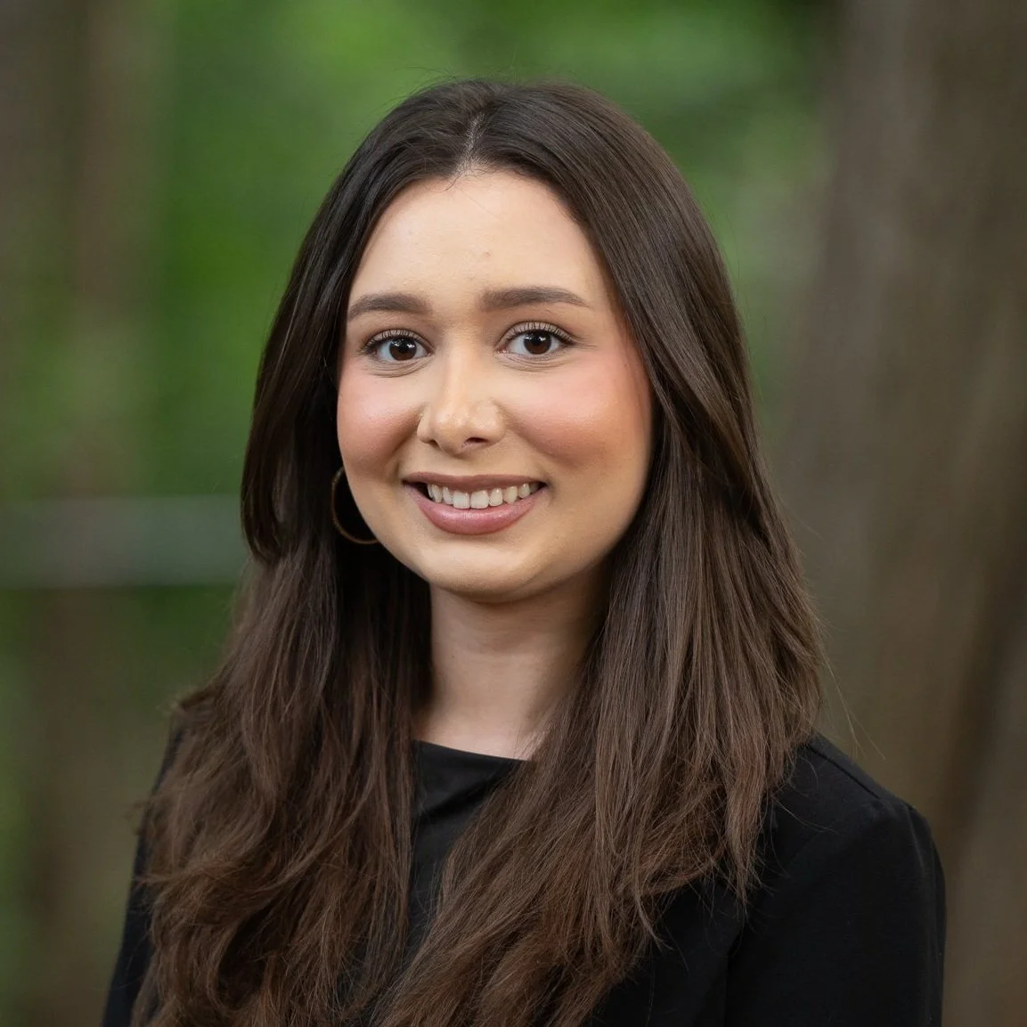 A young woman with long brown hair, wearing hoop earrings and a black top, smiling outdoors with trees in the background.