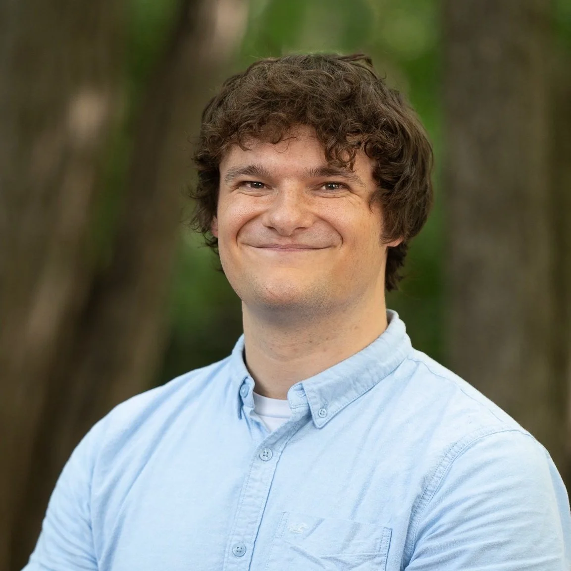 A young man with curly brown hair and a big smile, wearing a light blue button-up shirt, standing outdoors with blurred trees in the background.