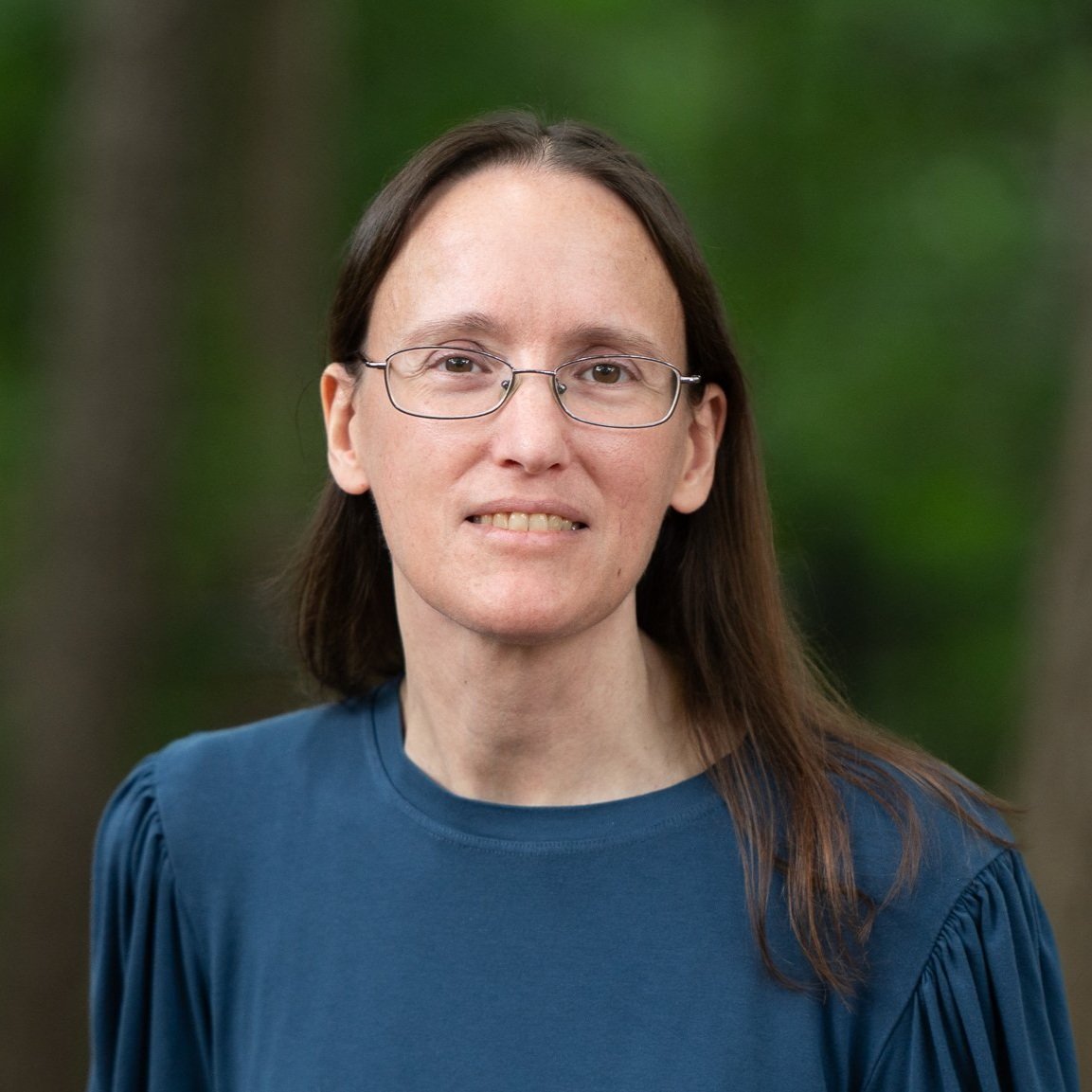 A woman with long brown hair, glasses, and a blue top standing outdoors with trees and greenery in the background.