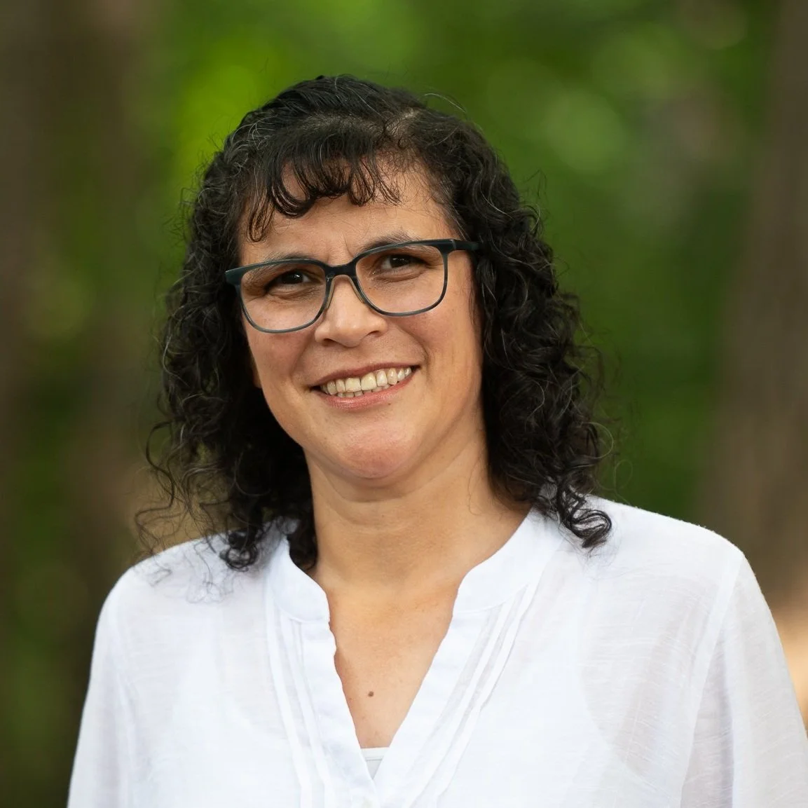 A smiling woman with curly dark hair and glasses, wearing a white blouse, standing outdoors with blurred green trees in the background.