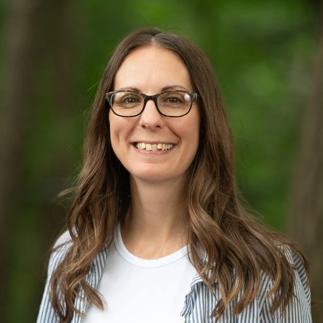 A woman with long wavy brown hair wearing glasses, a white shirt, and a striped jacket, smiling outdoors with a blurred green background.