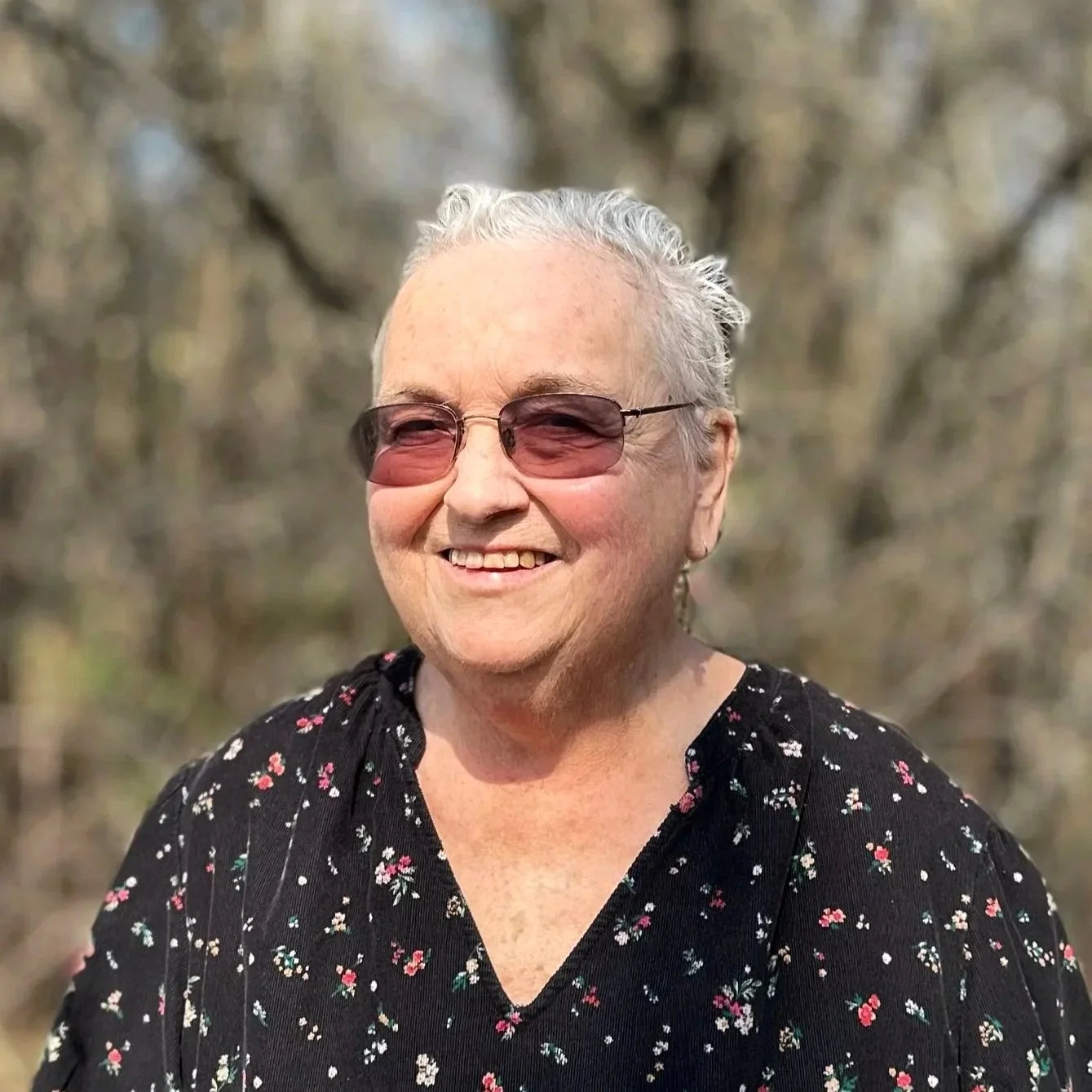 Smiling elderly woman with short gray hair, wearing sunglasses and a black floral blouse, outdoors with blurred trees in background.