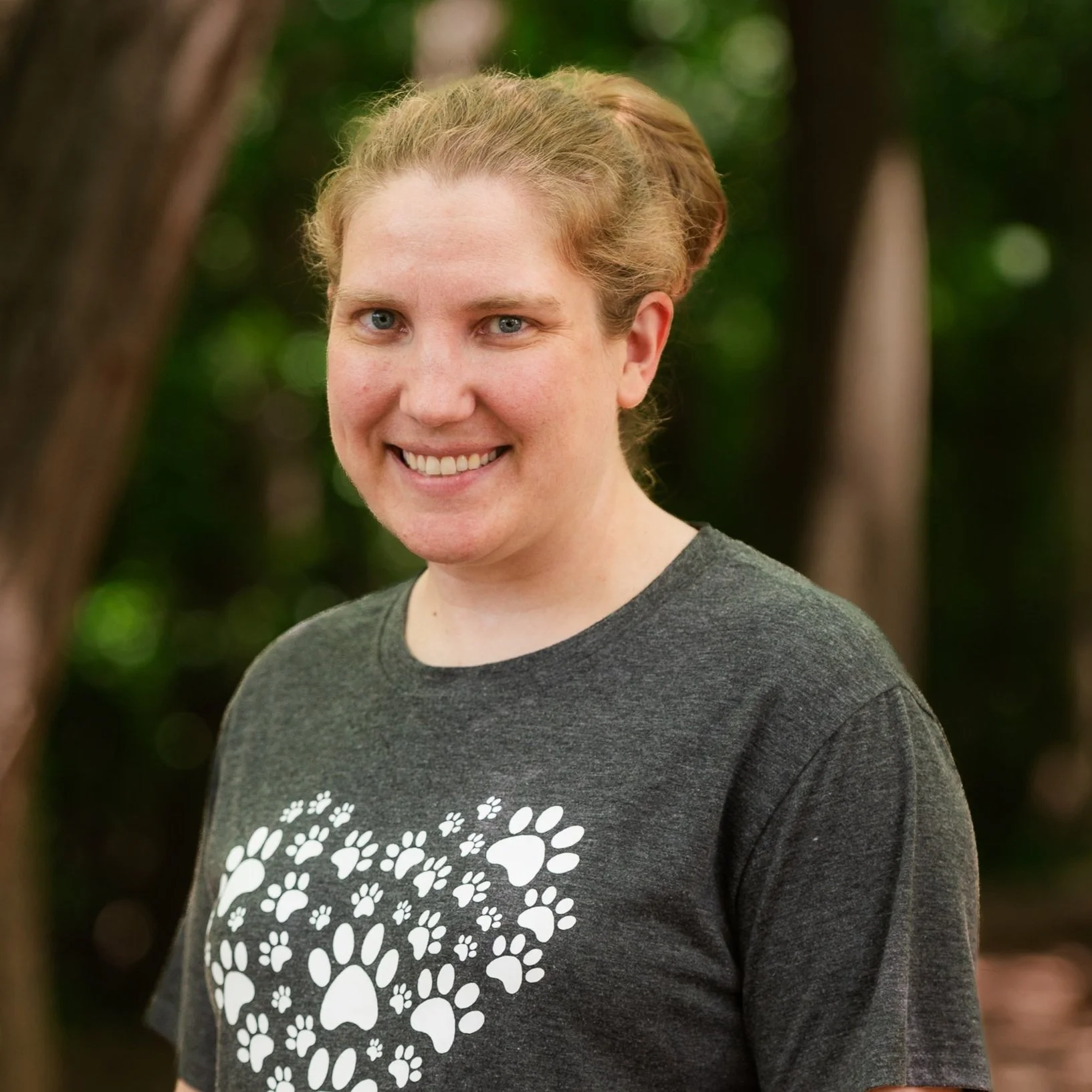 A smiling woman with blonde hair standing outdoors in front of trees, wearing a dark gray t-shirt with a heart design made of white paw prints.