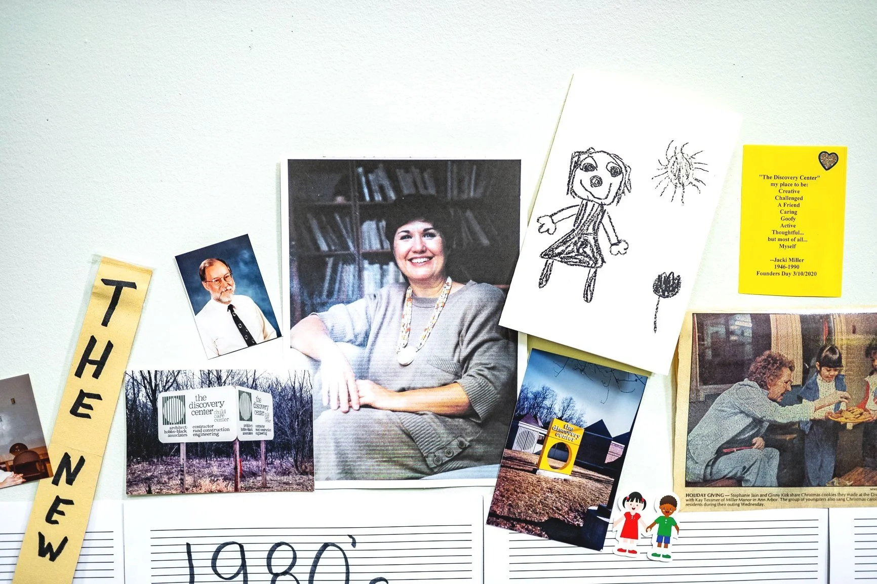 Collage of photos on a white wall, including a man and woman, children's drawings, a poem on a yellow card, and images of a building labeled "The Discovery Center". A strip of paper reads "THE NEW". A vintage holiday scene is also depicted.