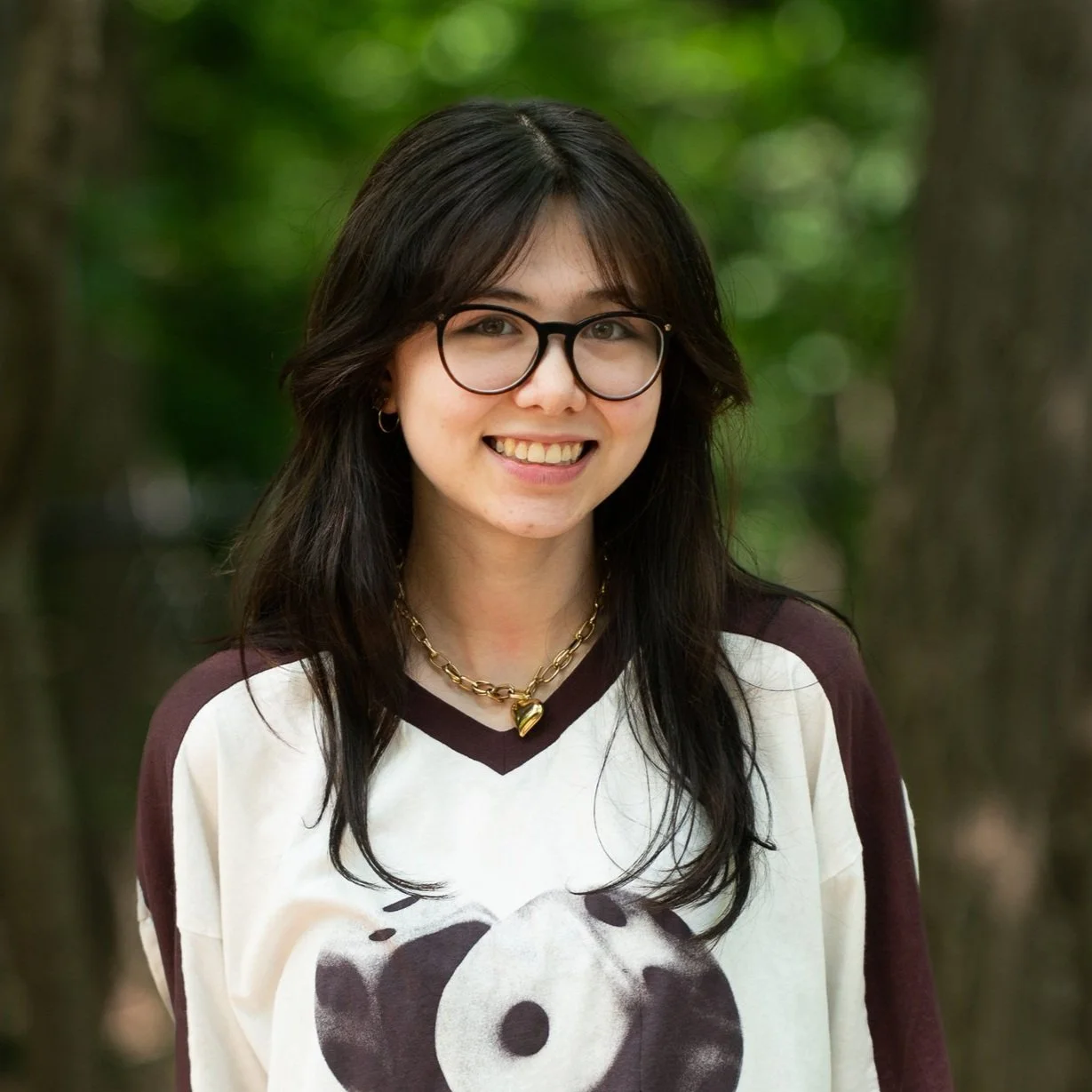 Young woman with dark hair, glasses, and gold jewelry smiling outdoors in front of trees.