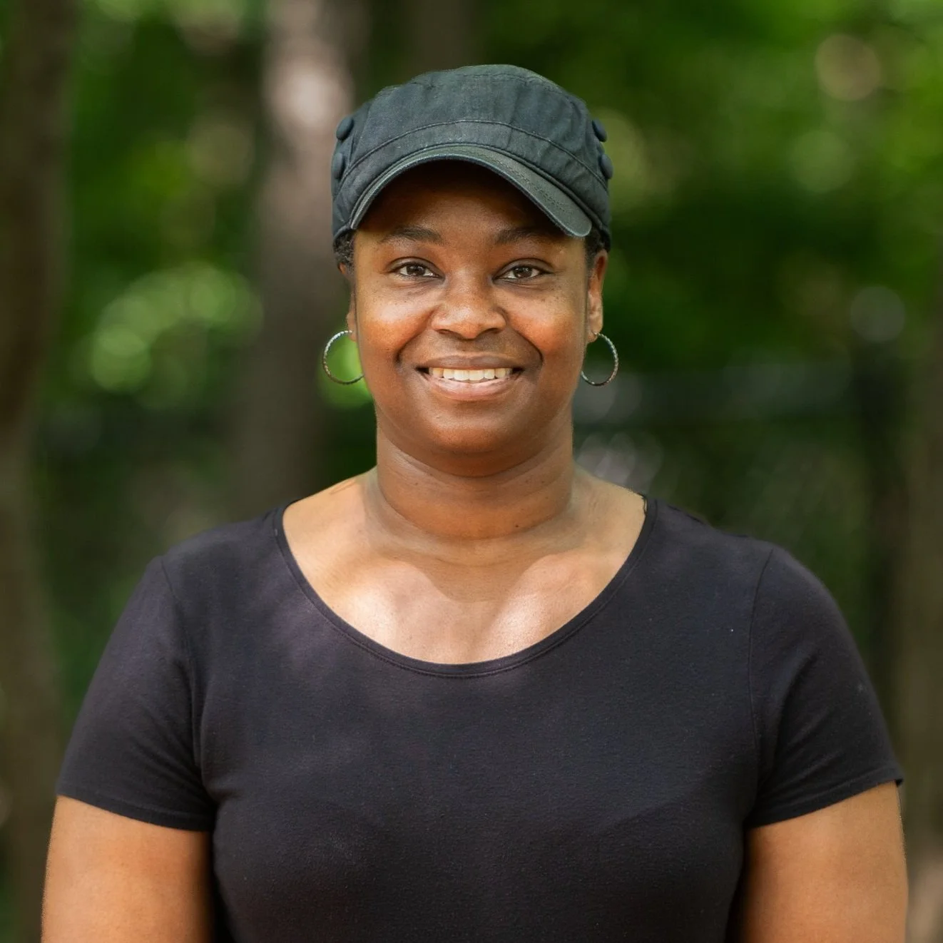 A smiling woman wearing a black cap, hoop earrings, and a black shirt, outdoors with green foliage in the background.