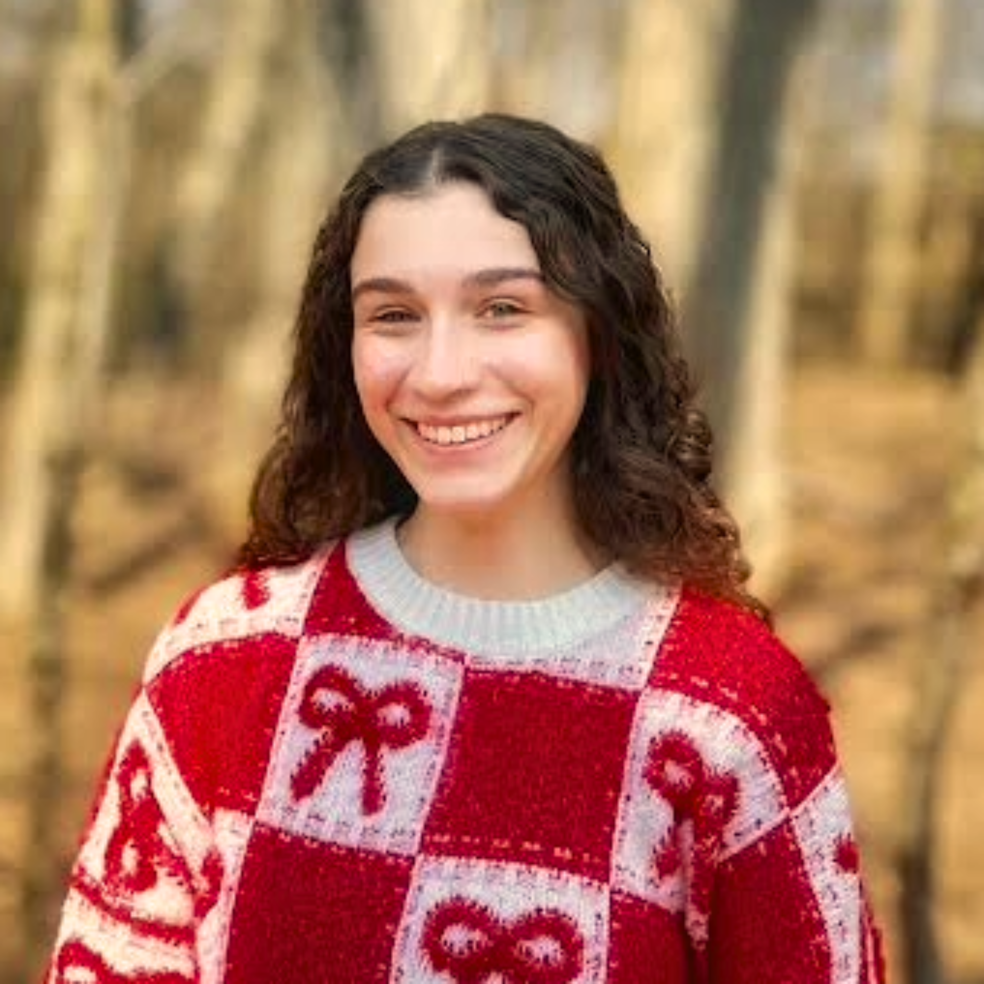 A woman with curly brown hair wearing a red and white Christmas sweater with a festive pattern, smiling outdoors with blurred trees in the background.