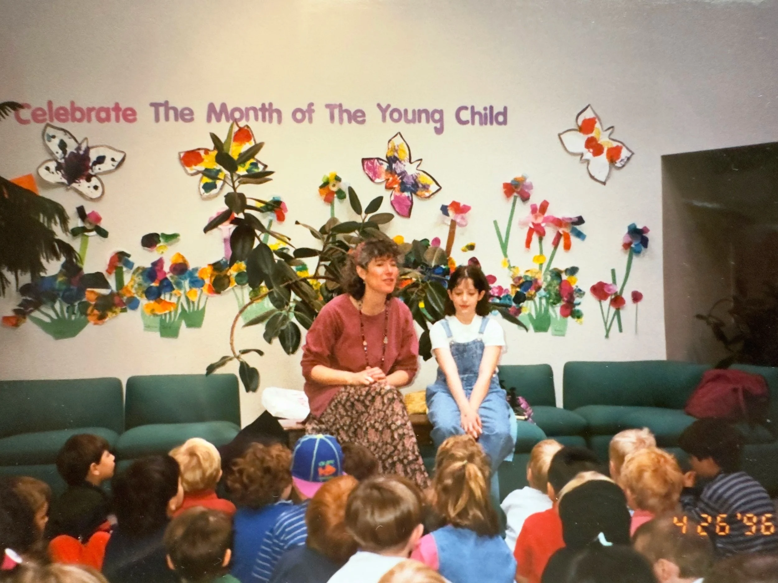 Two adults sitting with a group of children in a classroom decorated with colorful paper flowers and butterflies. A sign above reads "Celebrate The Month of The Young Child." The date on the image is April 26, 1996.