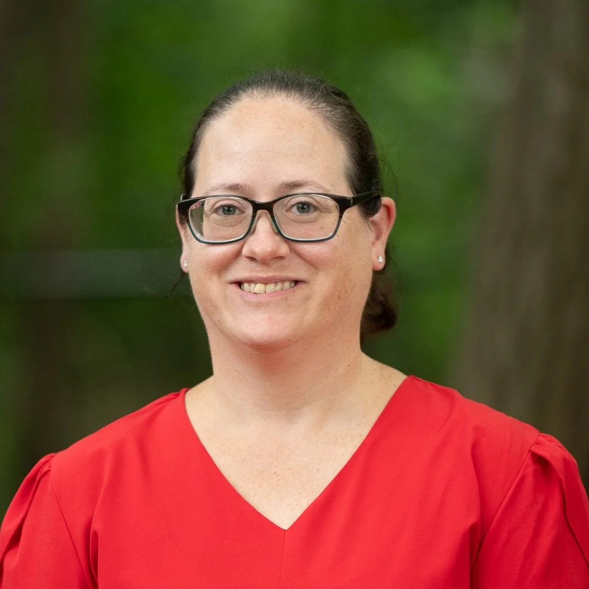 A woman with glasses wearing a red top, smiling, with a blurred green outdoor background.