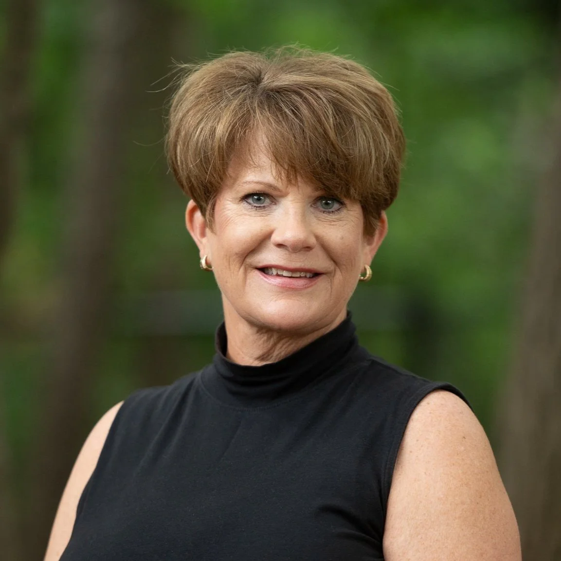 Portrait of a middle-aged woman with short brown hair, wearing a black sleeveless turtleneck, smiling outdoors with greenery in the background.
