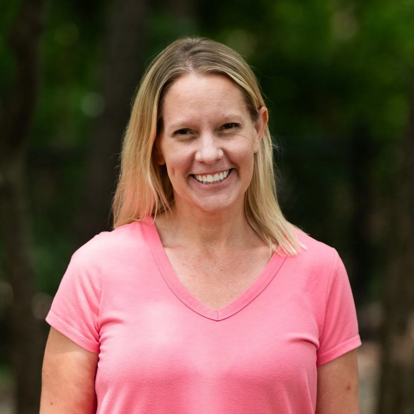 A woman with blonde hair smiling outdoors in a pink V-neck shirt, with a blurred green forest background.