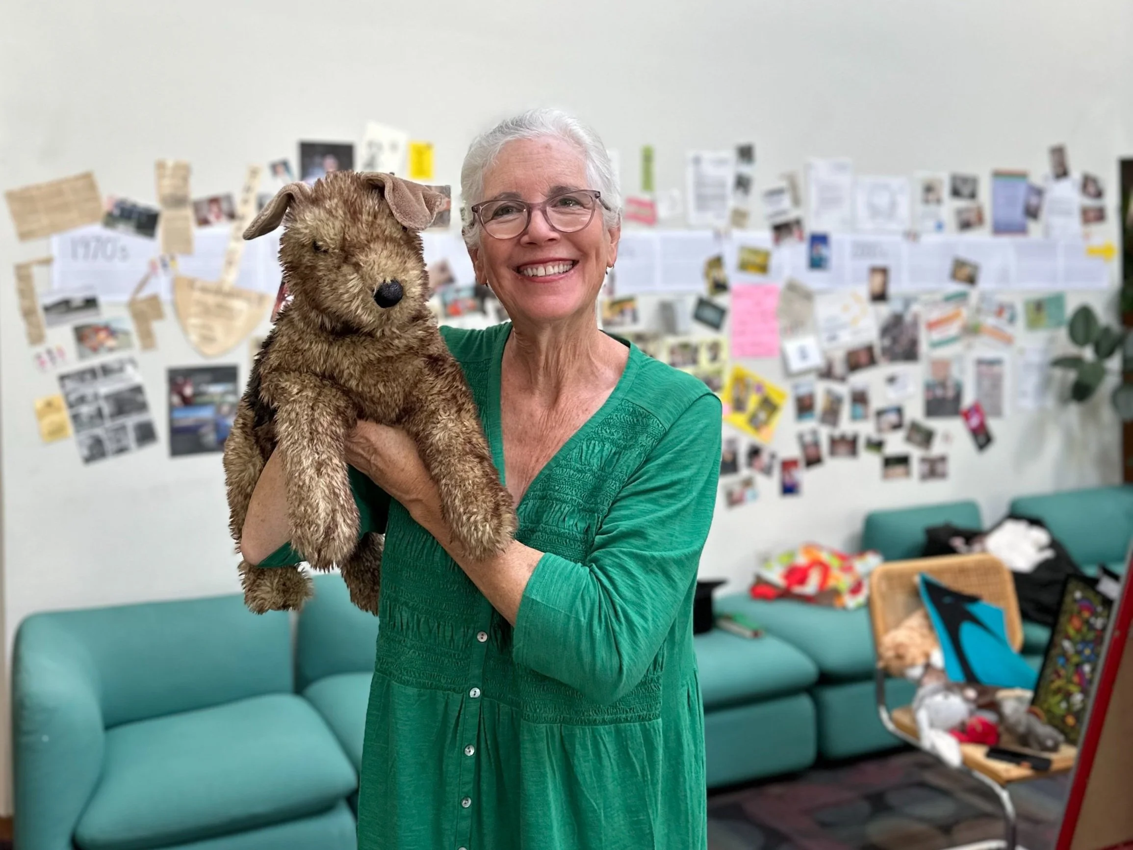 Person in a green shirt holding a plush dog toy, standing in a room with a collage of photos on the wall and green couches in the background.