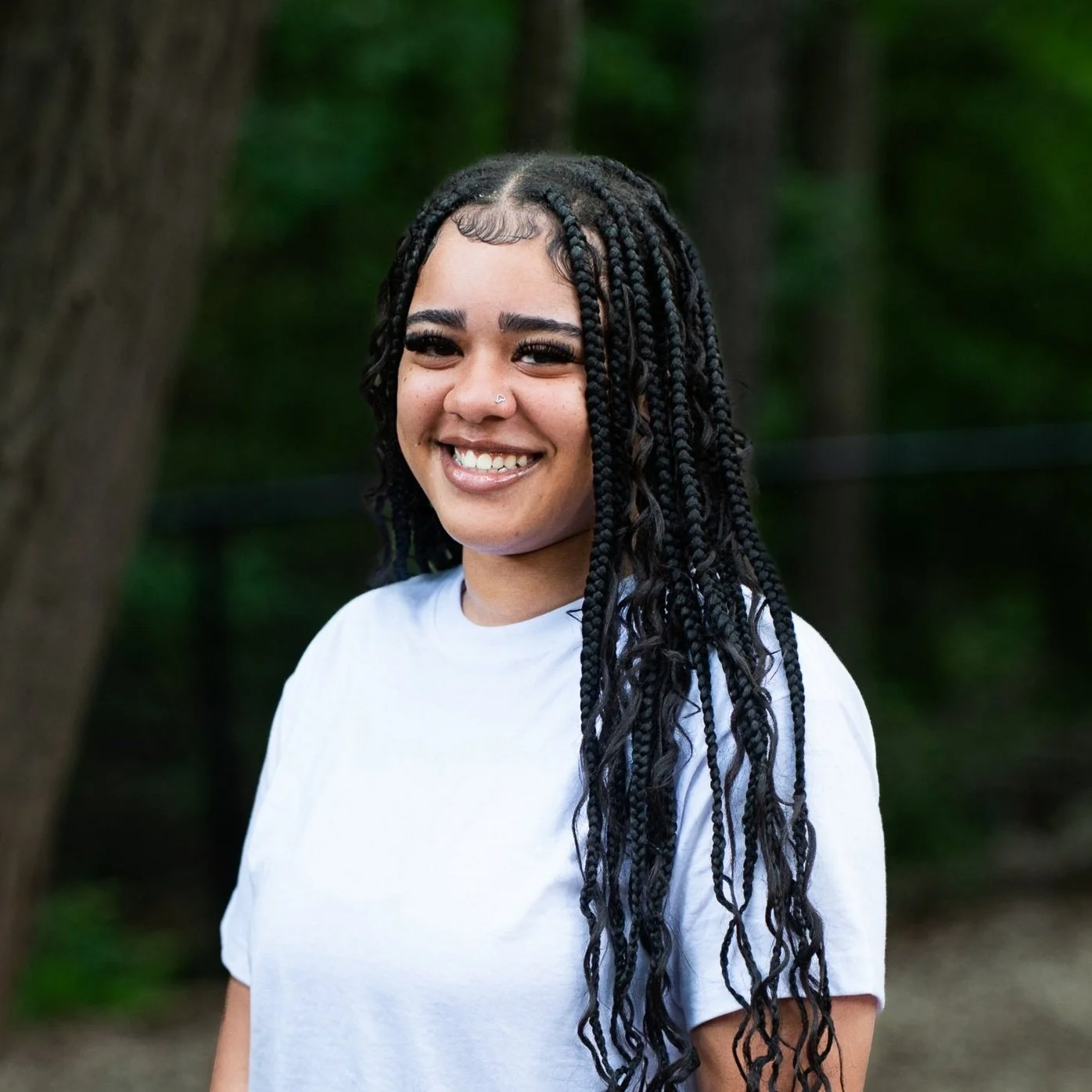 A young woman with dark, braided hair, smiling outdoors with a forest background.