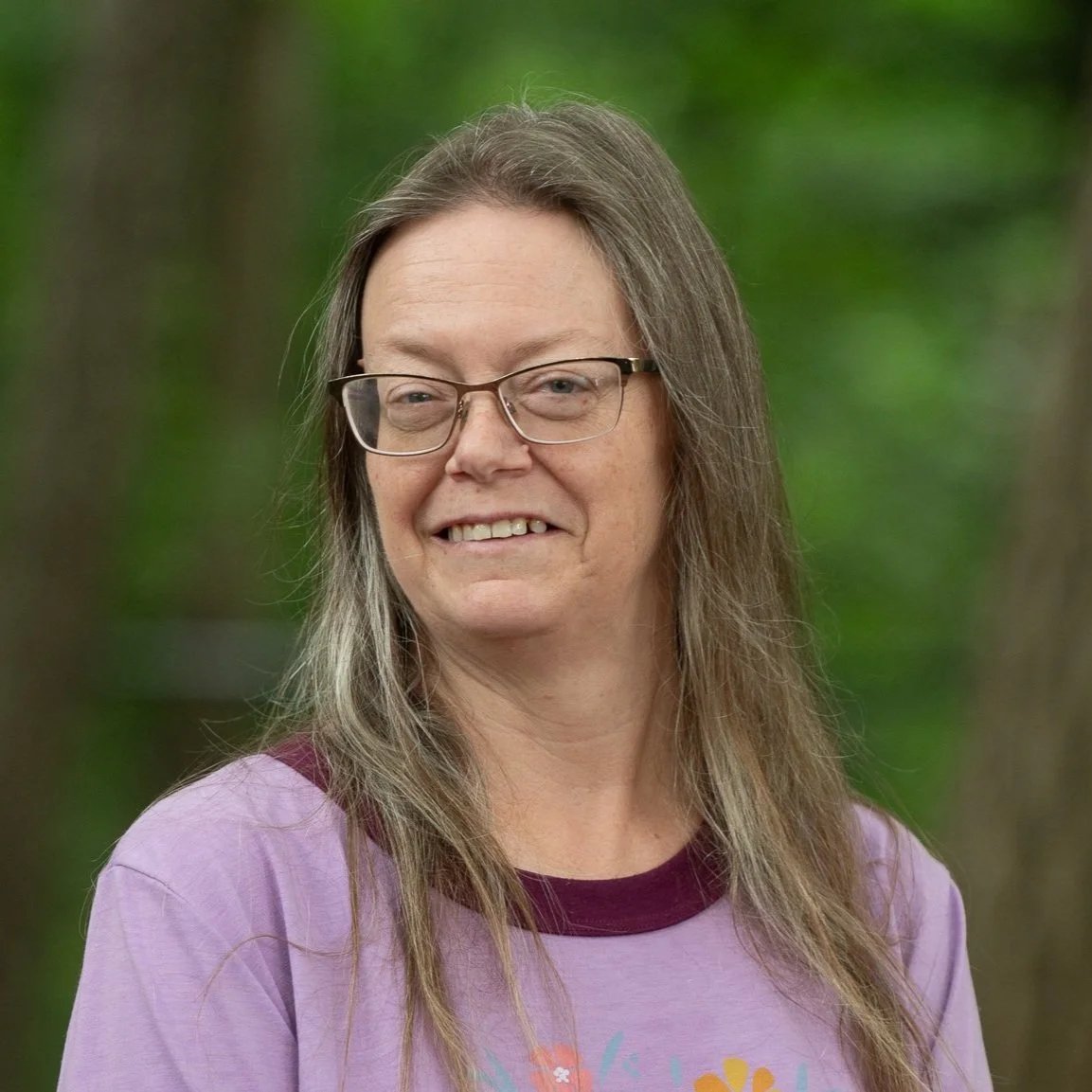 A woman with glasses and long brown hair smiling outdoors with green trees in the background.