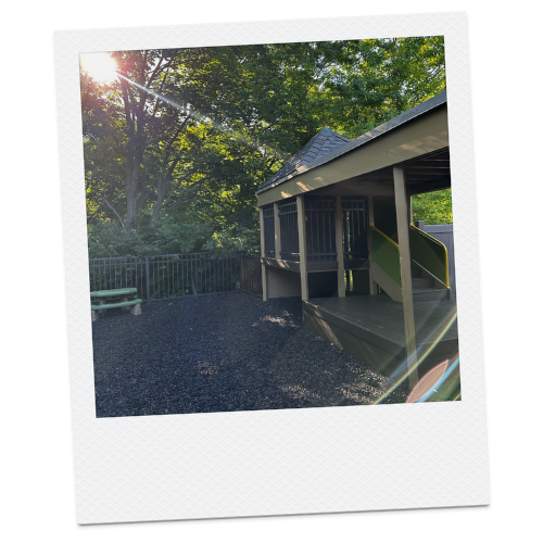 A playground with a wooden structure featuring a slide, surrounded by trees and sunlight streaming through the leaves.