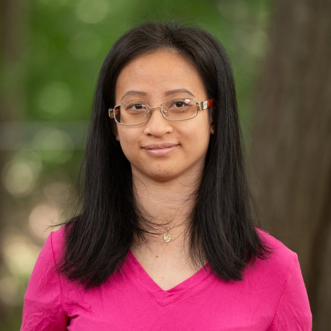 A young woman with long black hair, wearing glasses, a pink shirt, and a gold necklace, stands outdoors with a blurred green background.