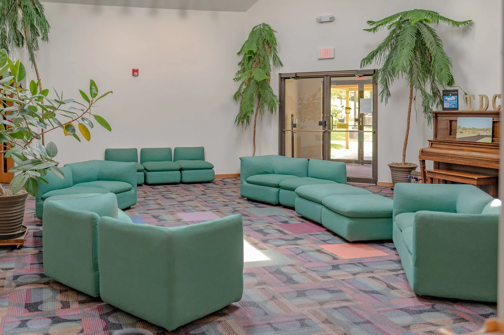 A waiting room with green upholstered chairs arranged in a semi-circle, potted plants, and a painted piano.