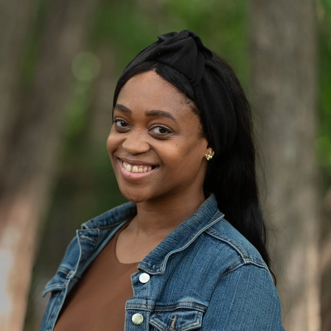 A woman smiling outdoors with a blurred forest background, wearing a black headwrap, earrings, a brown top, and a blue denim jacket.