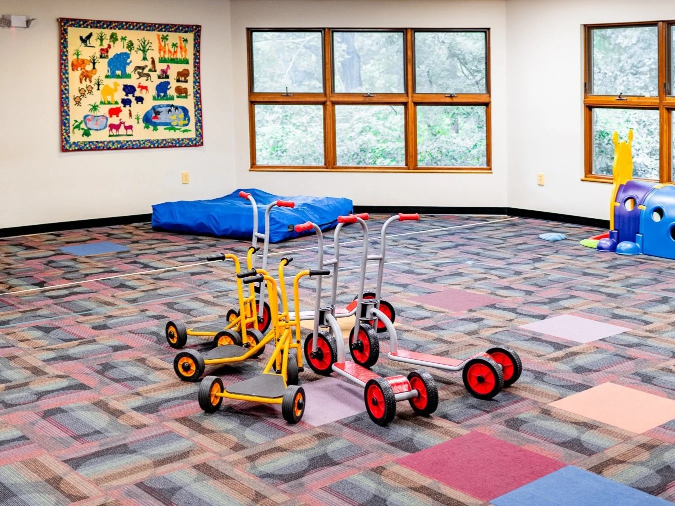 Indoor playroom with colorful carpet, push scooters, wall art, windows, and children's play equipment.