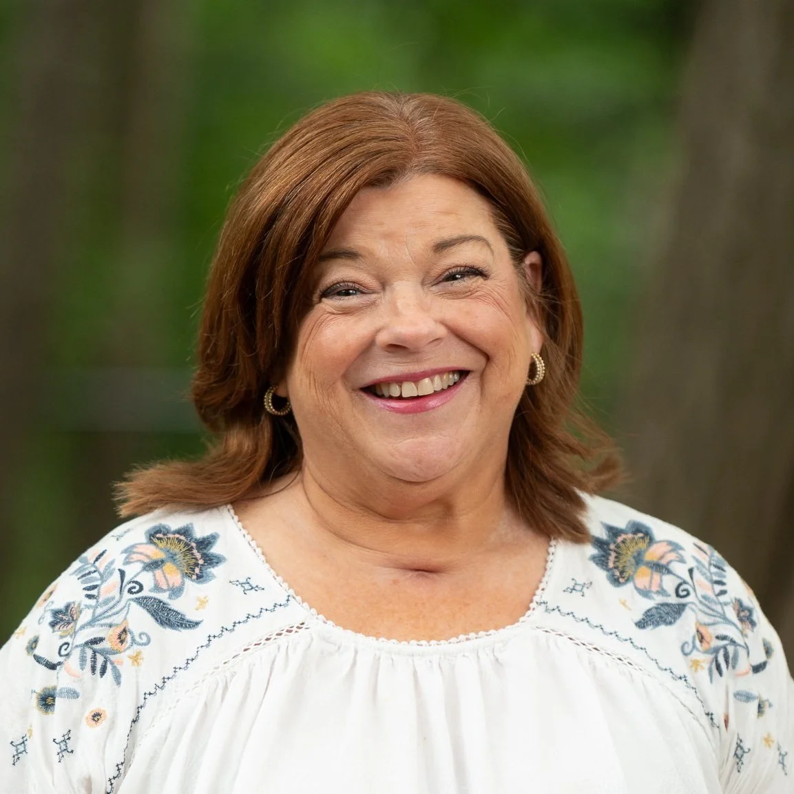 A woman with shoulder-length red hair smiling outdoors, wearing a white embroidered blouse and hoop earrings.