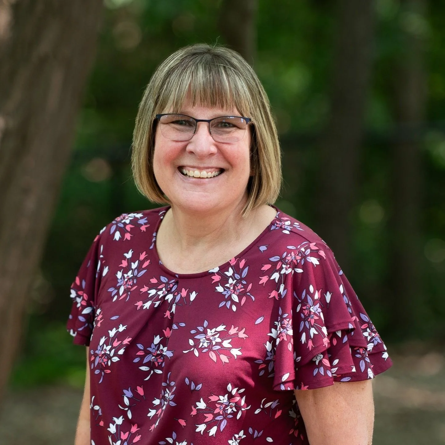A smiling woman with short blonde hair, glasses, and a maroon floral blouse standing outdoors in a wooded area.