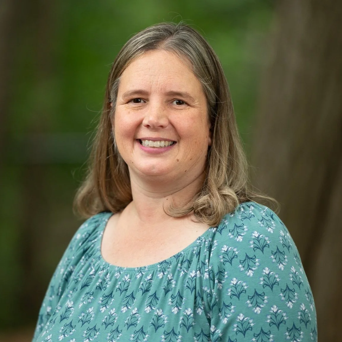 A woman with shoulder-length light brown hair, wearing a teal blouse with a floral pattern, smiling outdoors with a blurred green background.