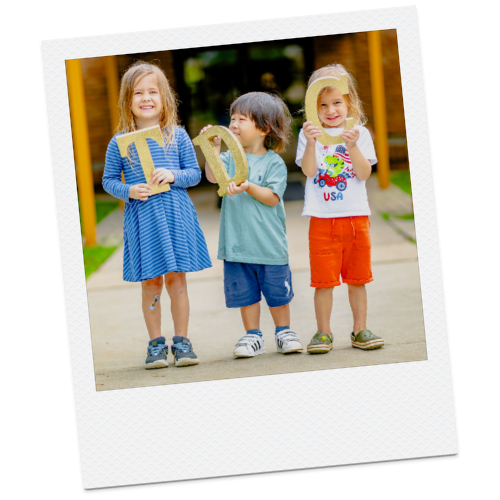Three children standing outdoors holding large numbers, with the girl on the left holding '1', the boy in the middle holding '0', and the girl on the right holding '2'.