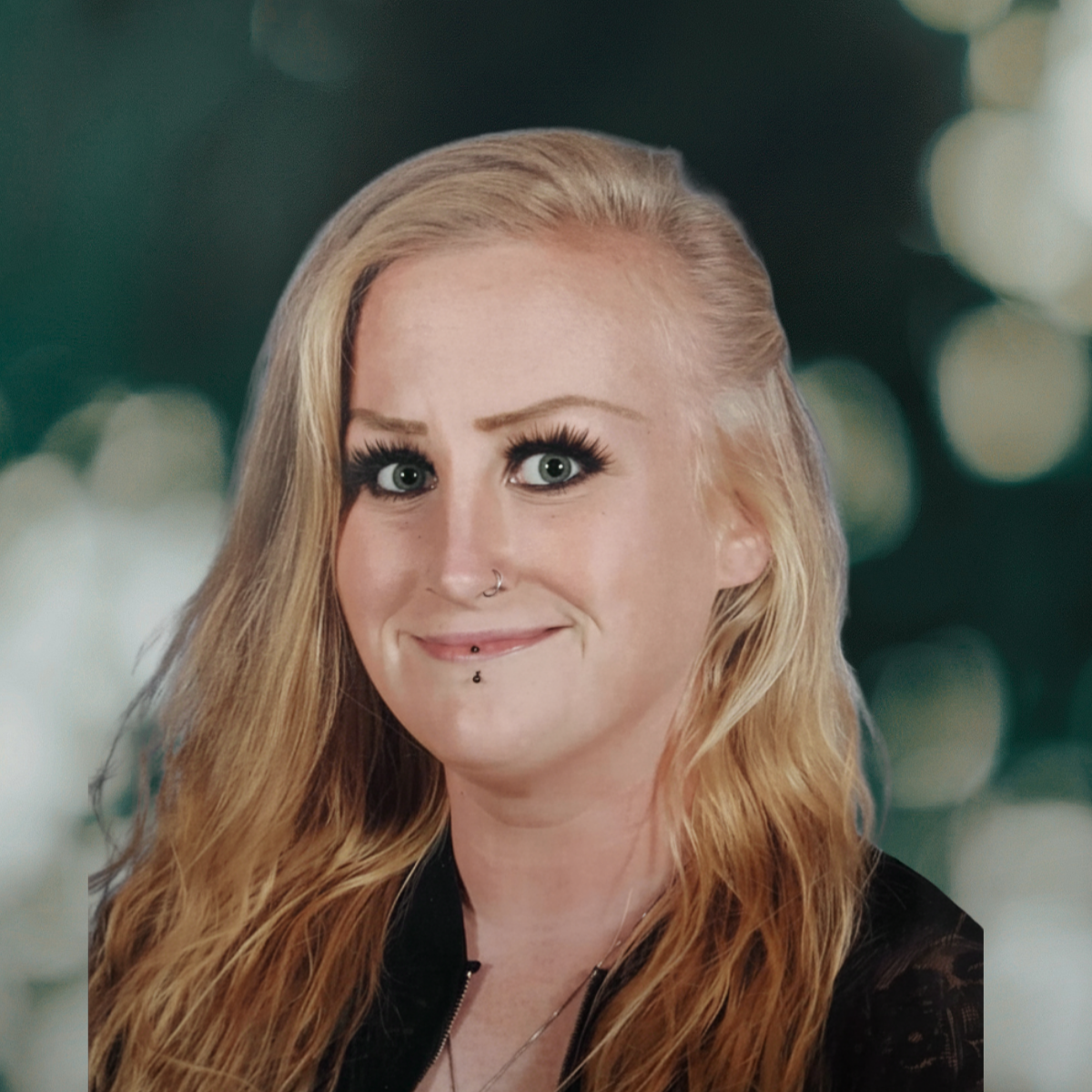 A woman with long, wavy blonde hair, blue eyes, and multiple facial piercings, smiling at the camera, with a blurred background of lights.