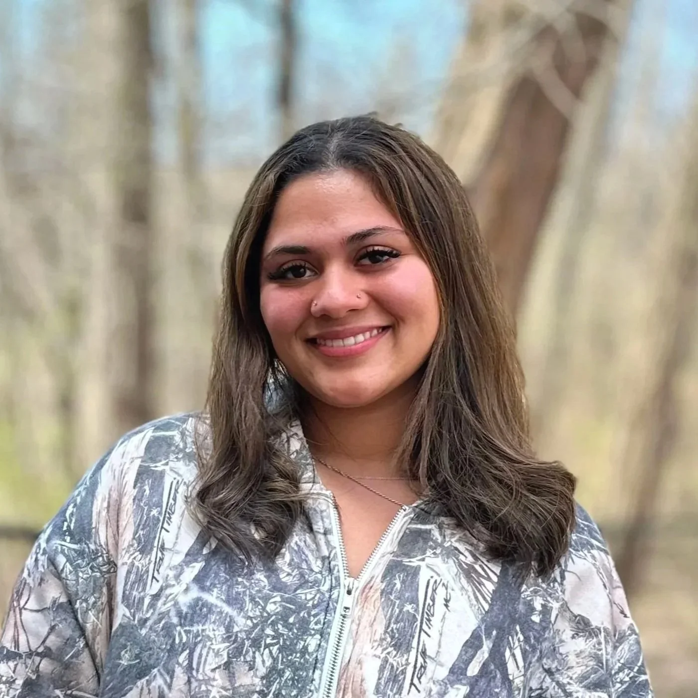 A young woman with long brown hair, smiling, standing outdoors in a wooded area with blurred trees in the background.